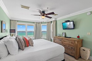 A bedroom with a bed, a television mounted on the wall, a ceiling fan, and a view of the ocean through large windows.