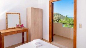 Un dormitorio con escritorio de madera, armario, cama con toalla blanca y balcón con vistas a zonas verdes y colinas.