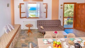 Un salón acogedor con un sofá, una mesa de comedor con platos y frutas, una mesita redonda con un plato, y vista al mar a través de una ventana.