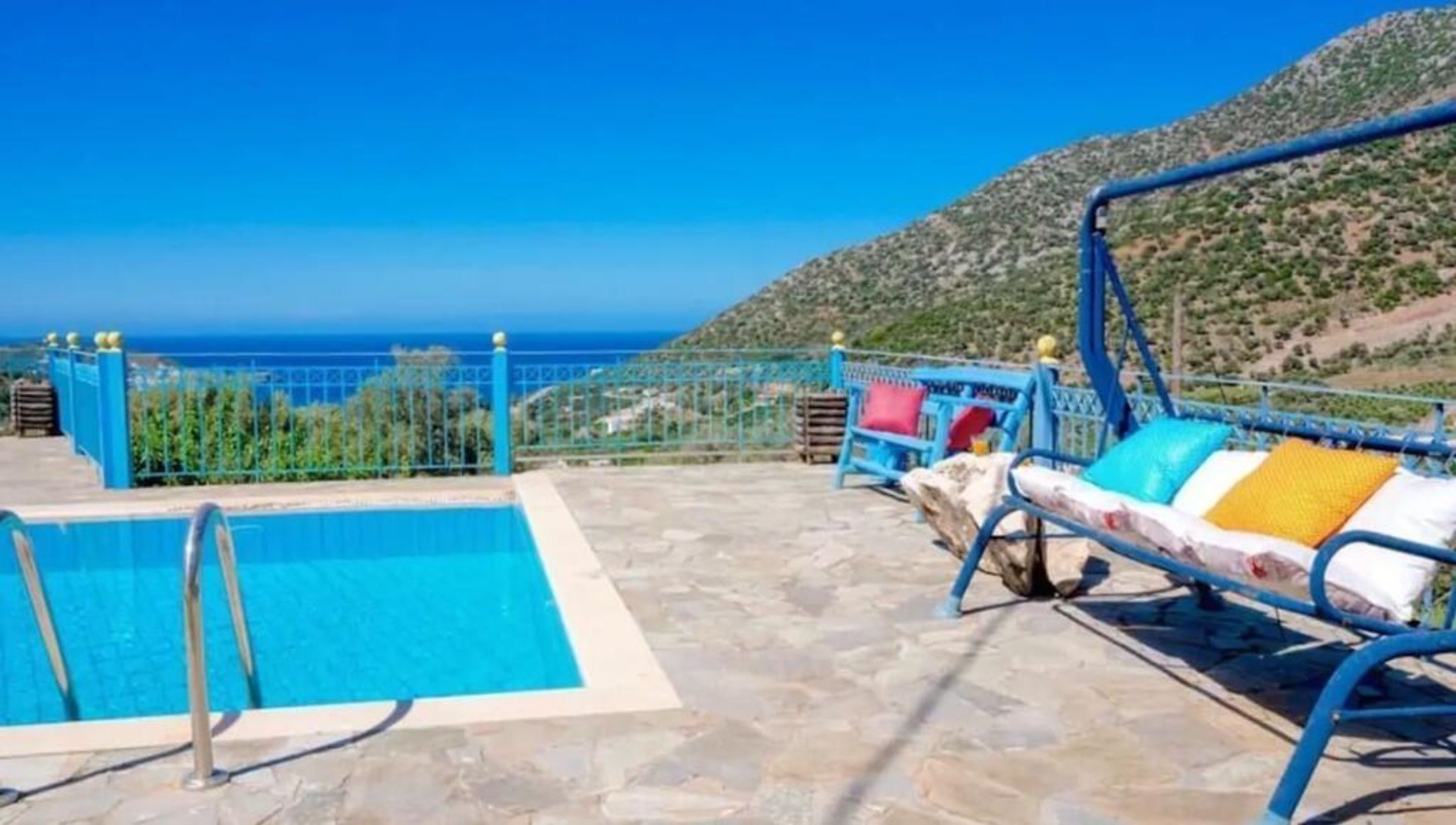 Un espace piscine avec vue sur la mer, une chaise longue avec des coussins et une clôture en métal.