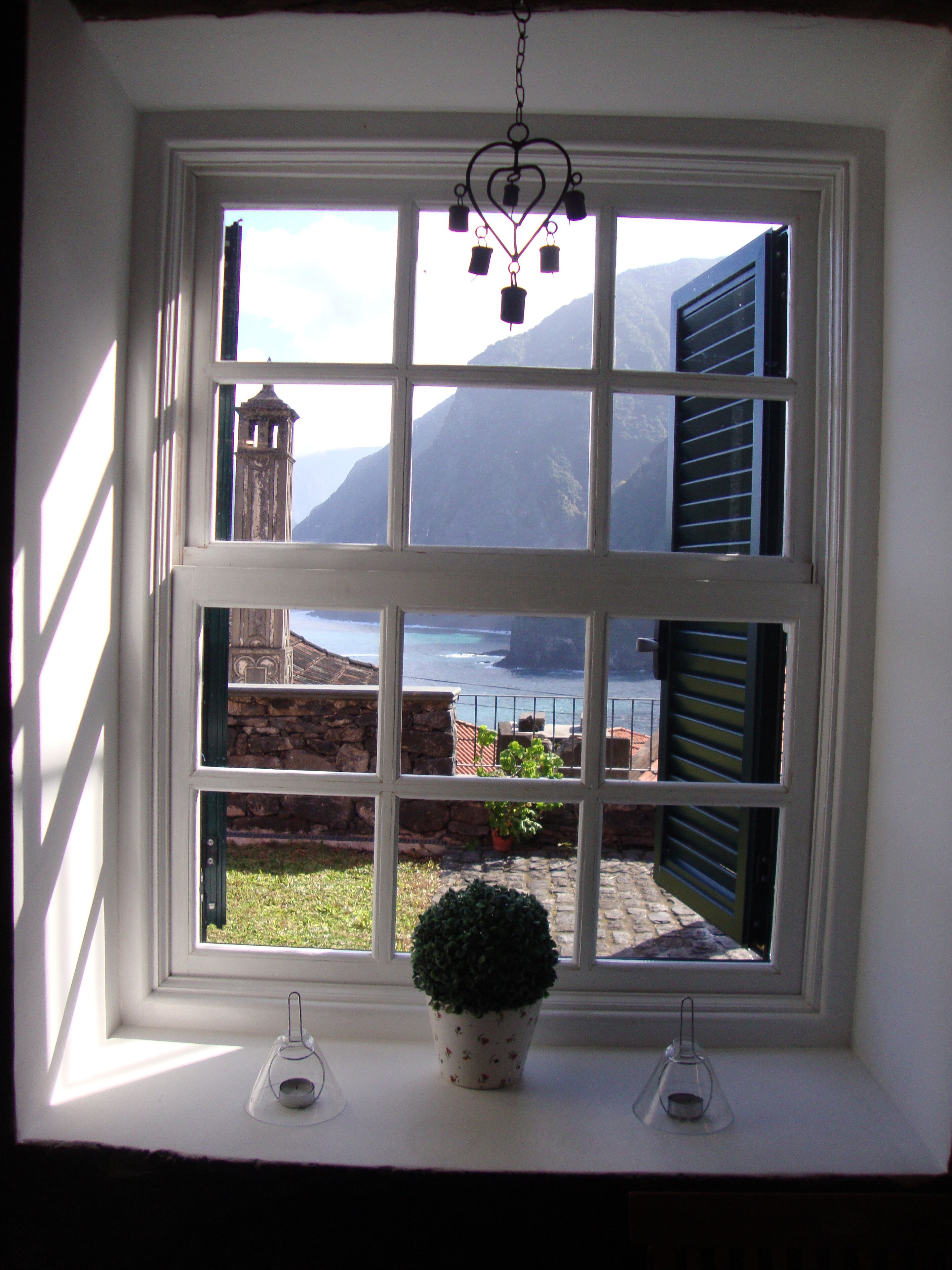 A window with a view of a coastal landscape, including a lighthouse and a building with shutters.
