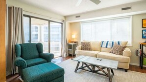 Een woonkamer met een groene fauteuil, een beige bank met kussens met dessin, een salontafel, en uitzicht op een balkon door grote ramen.