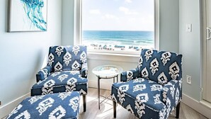 A room with two patterned armchairs, a small round table, and a large window overlooking the beach.