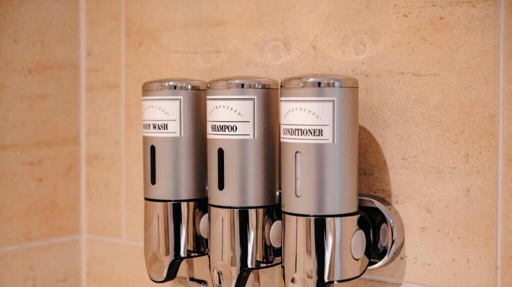 Drei metallische Spender mit Beschriftung für Duschgel, Shampoo und Spülung an einer gefliesten Wand angebracht.