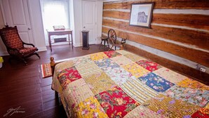 A bedroom with a wooden log wall, a quilted bedspread, a rocking chair, a small table with a book, and a window with curtains. - Fulkerson-Hilton historic homestead (Hiltons)