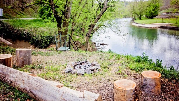 A riverside campsite with a fire pit, wooden stumps, and a metal railing. - Fulkerson-Hilton historic homestead (Hiltons)
