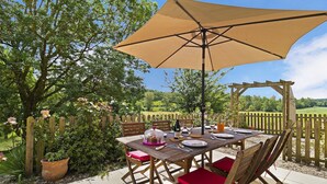An outdoor wooden table with a beige umbrella, surrounded by red cushioned chairs, set on a patio with a wooden fence and greenery in the background. - La Maison de Campagne - Le Farat (Auvillar)