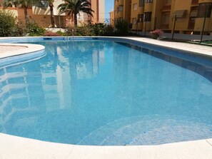 Ein klarer Pool mit gefliesten Rändern, umgeben von einer Betonplattform und Palmen, mit einem Gebäude im Hintergrund.