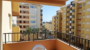 Un balcó amb una cadira de plàstic blanca i una taula, amb vista a un carrer amb palmeres i edificis d'altes plantes.