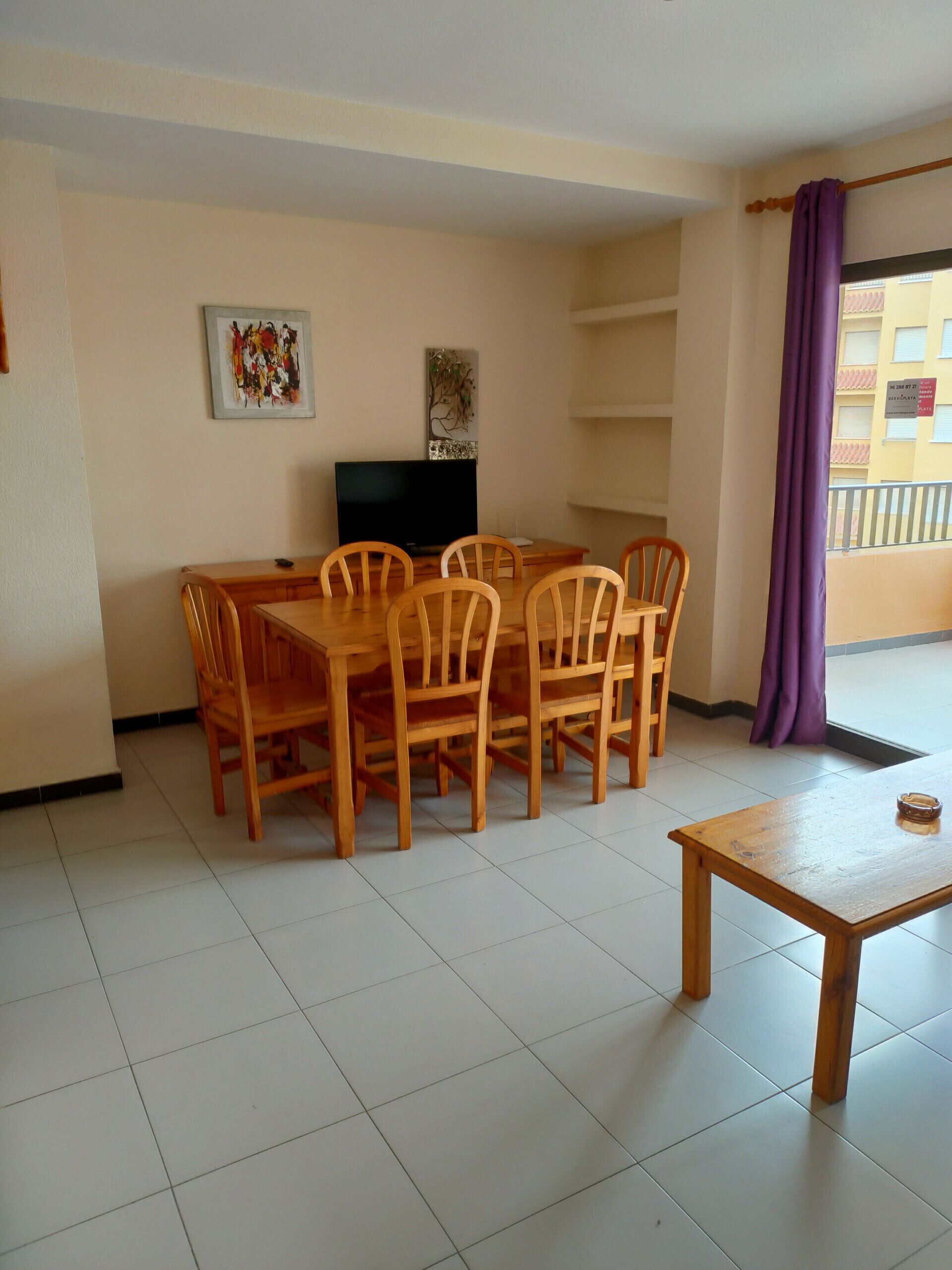 Un comedor con una mesa y sillas de madera, un televisor y un balcón con cortina morada.