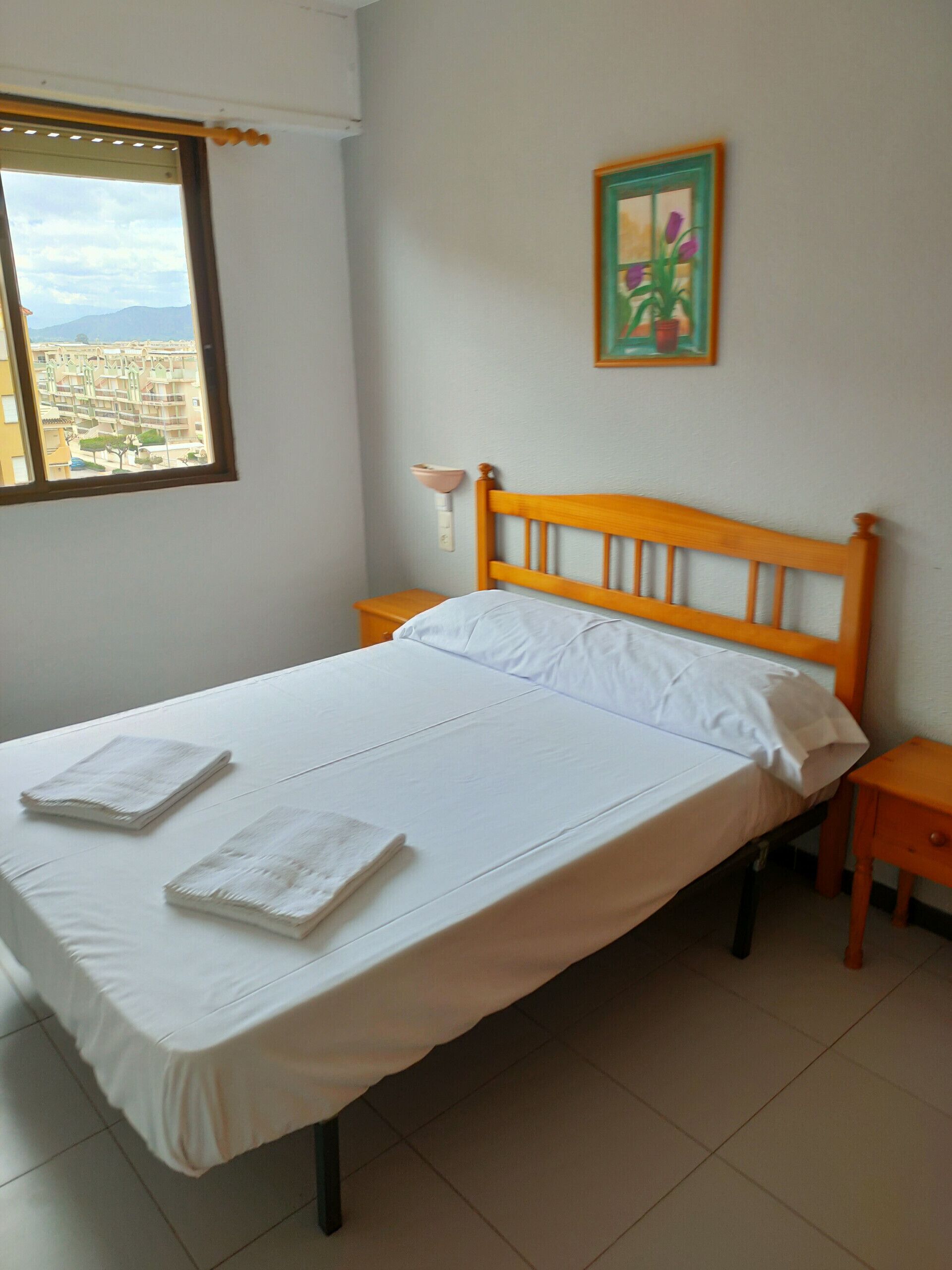A single-bed room with a wooden headboard, white bedding, a small bedside table, a wall-mounted nightstand, and a window with a view of buildings.