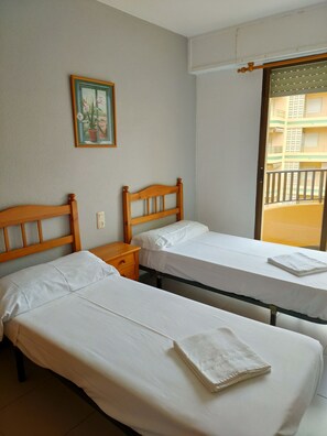 Une chambre d’hôtel avec deux lits simples, une tête de lit en bois, une table de chevet, un tableau au mur et un balcon avec vue sur un immeuble.