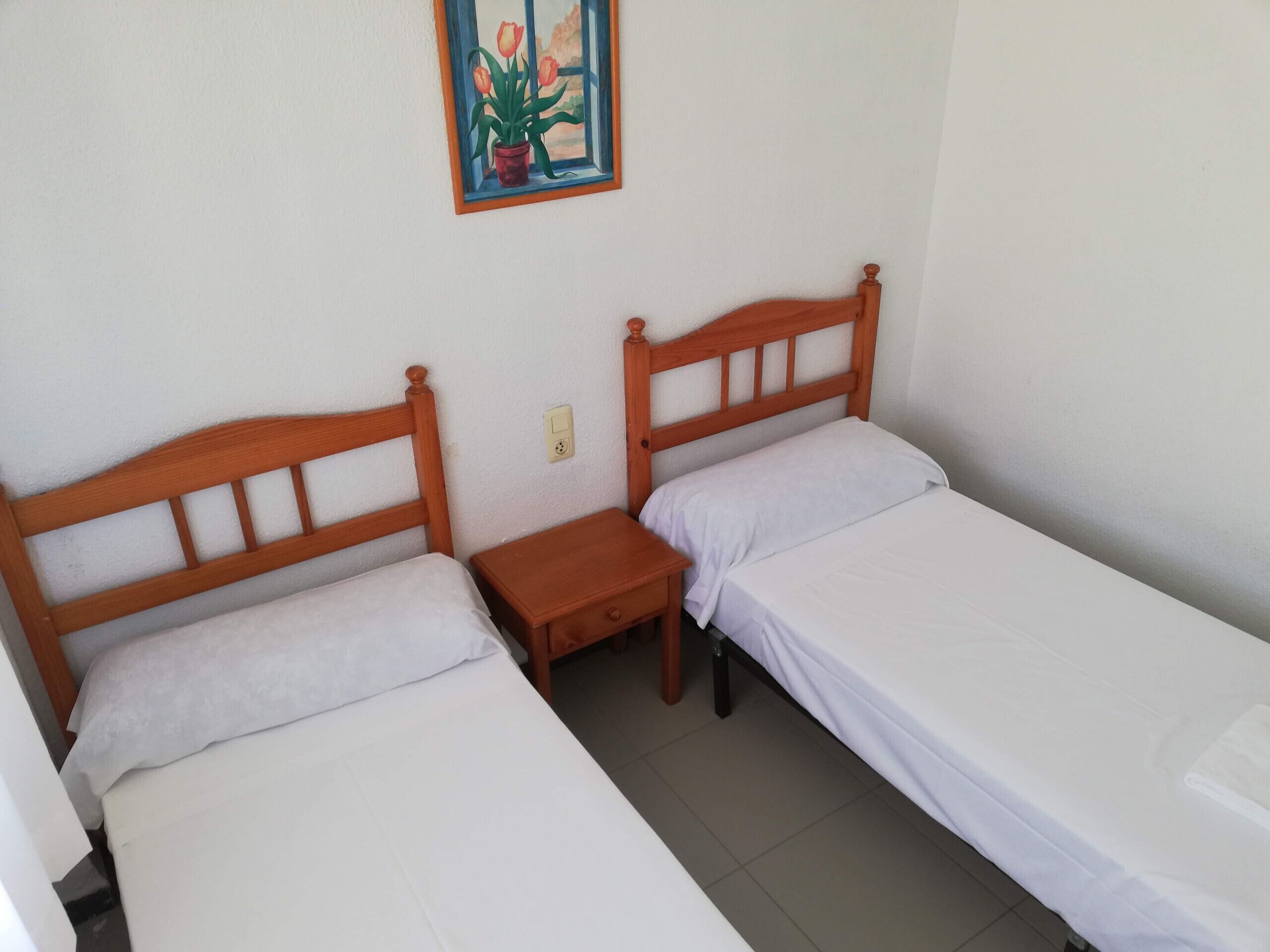 Una habitación pequeña y limpia con dos camas individuales, una mesita de noche y un cuadro de flores colgado en la pared.