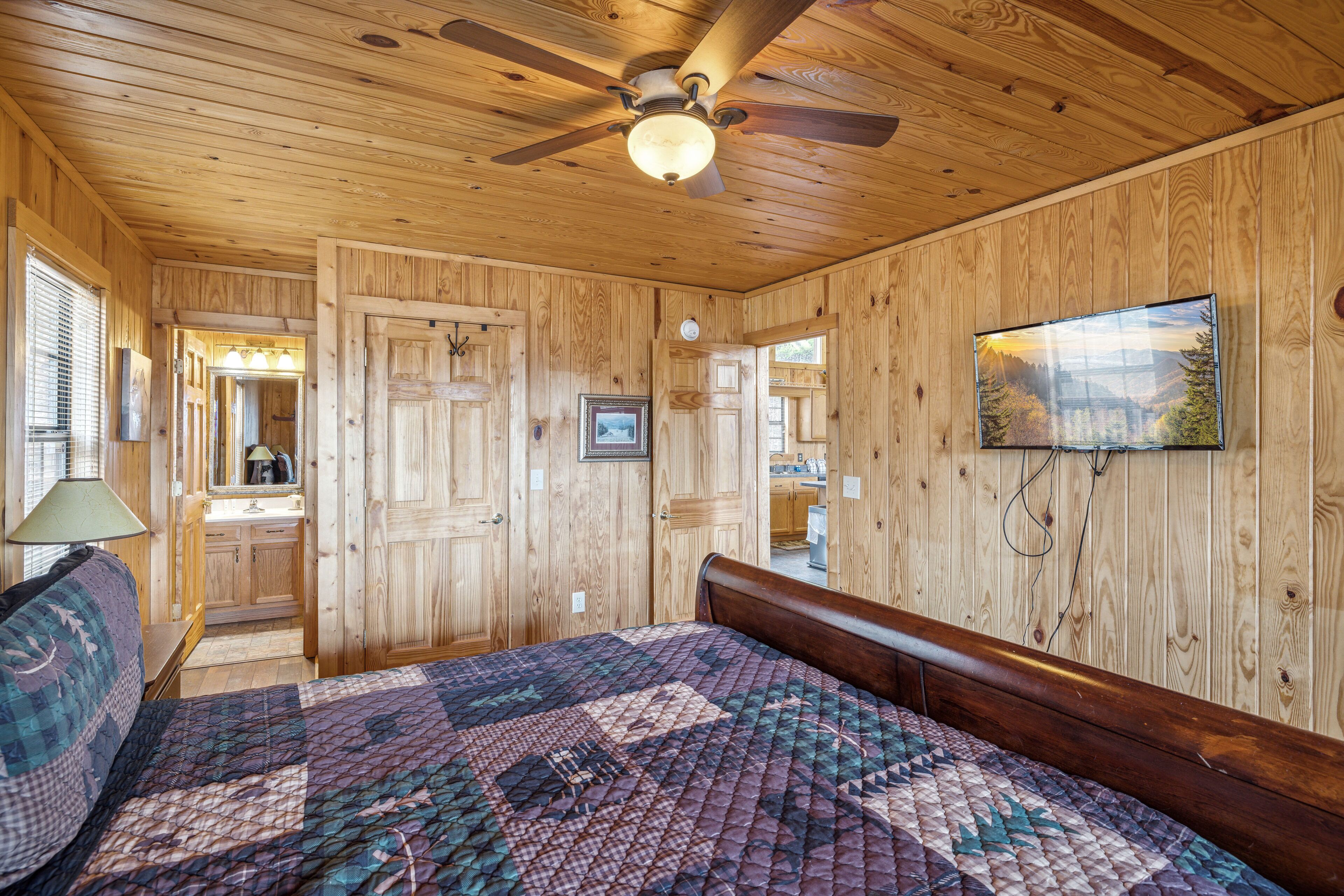 A wooden-paneled bedroom with a bed, a ceiling fan, a TV, and a bathroom visible through an open door.