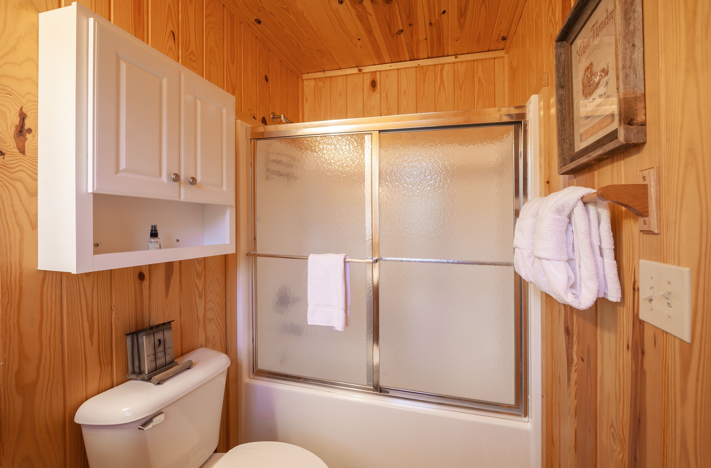 Une salle de bain aux murs lambrissés, équipée de toilettes, d’une douche en verre et d’un meuble blanc placé au-dessus des toilettes.