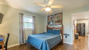 Une chambre à coucher comprenant un lit, un ventilateur de plafond, une fenêtre avec des rideaux et une porte donnant sur la cuisine.