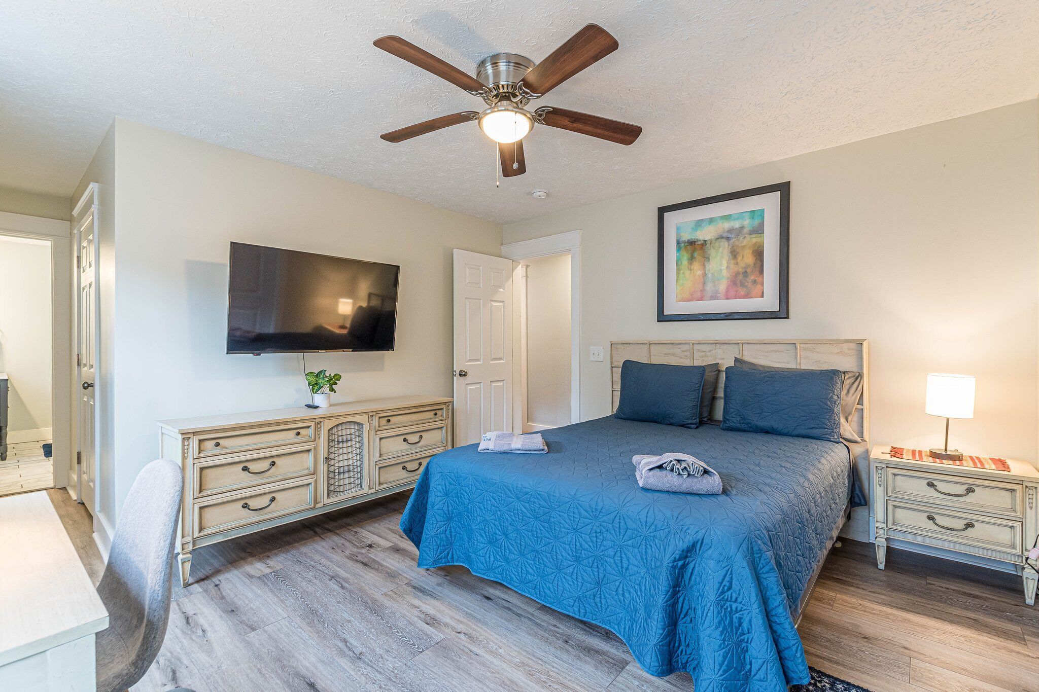 A bedroom with a bed, a ceiling fan, a TV, a painting, and a nightstand.