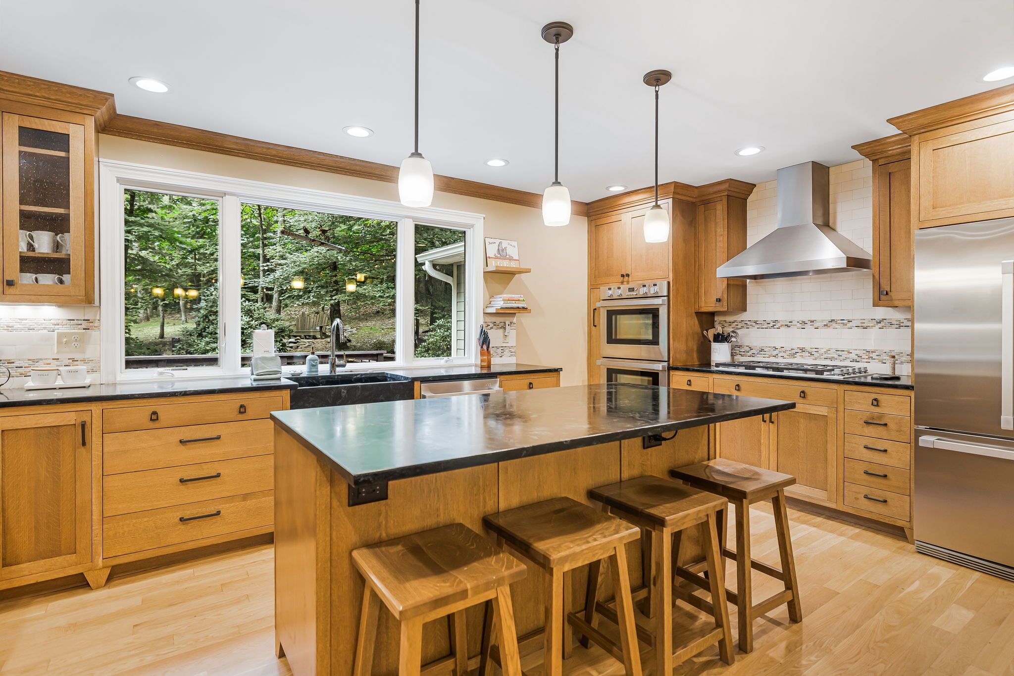 A modern kitchen with wooden cabinets, a large island with stools, stainless steel appliances, and a view of the outdoors.