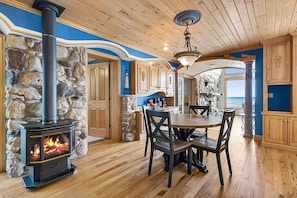 A room with a stone wall, a fireplace, a dining table with chairs, and a view of the ocean. - Door County Area Waterfront Estate | Game Room (Algoma)