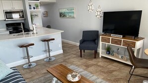 Una cocina moderna con microondas, estufa y taburetes de bar. Se observa un sillón gris y un televisor sobre un mueble de madera.