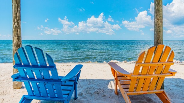 Dos sillas Adirondack sobre una playa arenosa, con un cielo azul despejado y un mar tranquilo al fondo.