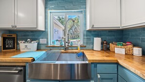 Una cocina con un lavamanos de acero inoxidable, una ventana con vistas a árboles y un cartel que dice 'Will cook for wine'.