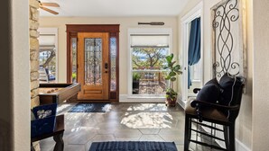 Un vestíbulo con puerta de madera, puerta de vidrio con reja de metal, un banco, una planta en maceta y vista al exterior.