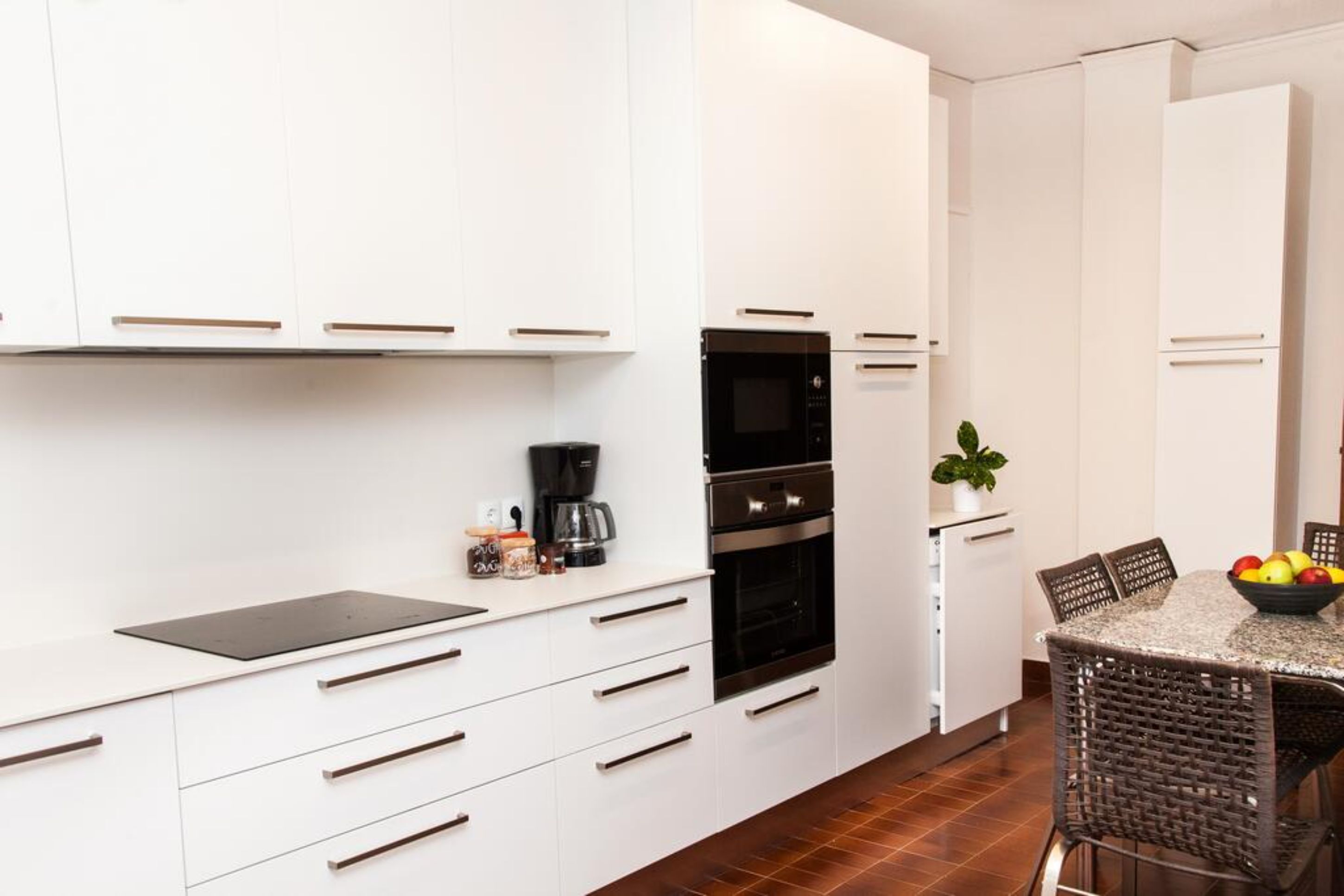 Een moderne keuken met witte kasten, een inbouwoven en een eethoek met rieten stoelen en een tafel.