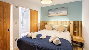 A hotel room with a bed, a bedside table, a nightstand, a wall-mounted lamp, a painting, and a shower area. - Hot Tub Haven with Dreamy Sea Vista! (Ilfracombe)