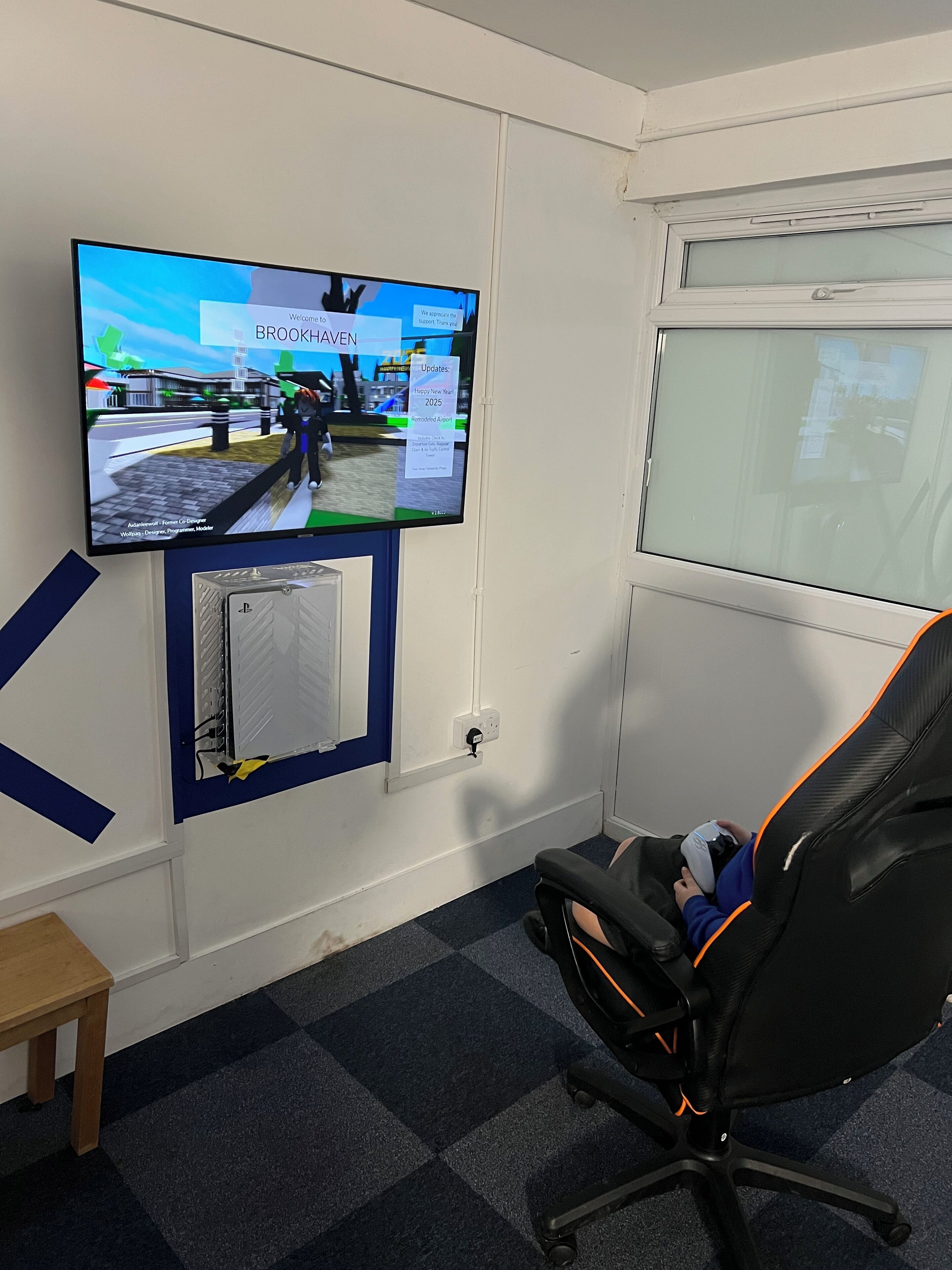 Quarto com televisão montada exibindo um ambiente virtual, uma cadeira gamer e uma janela.
