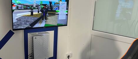 Quarto com televisão montada exibindo um ambiente virtual, uma cadeira gamer e uma janela.