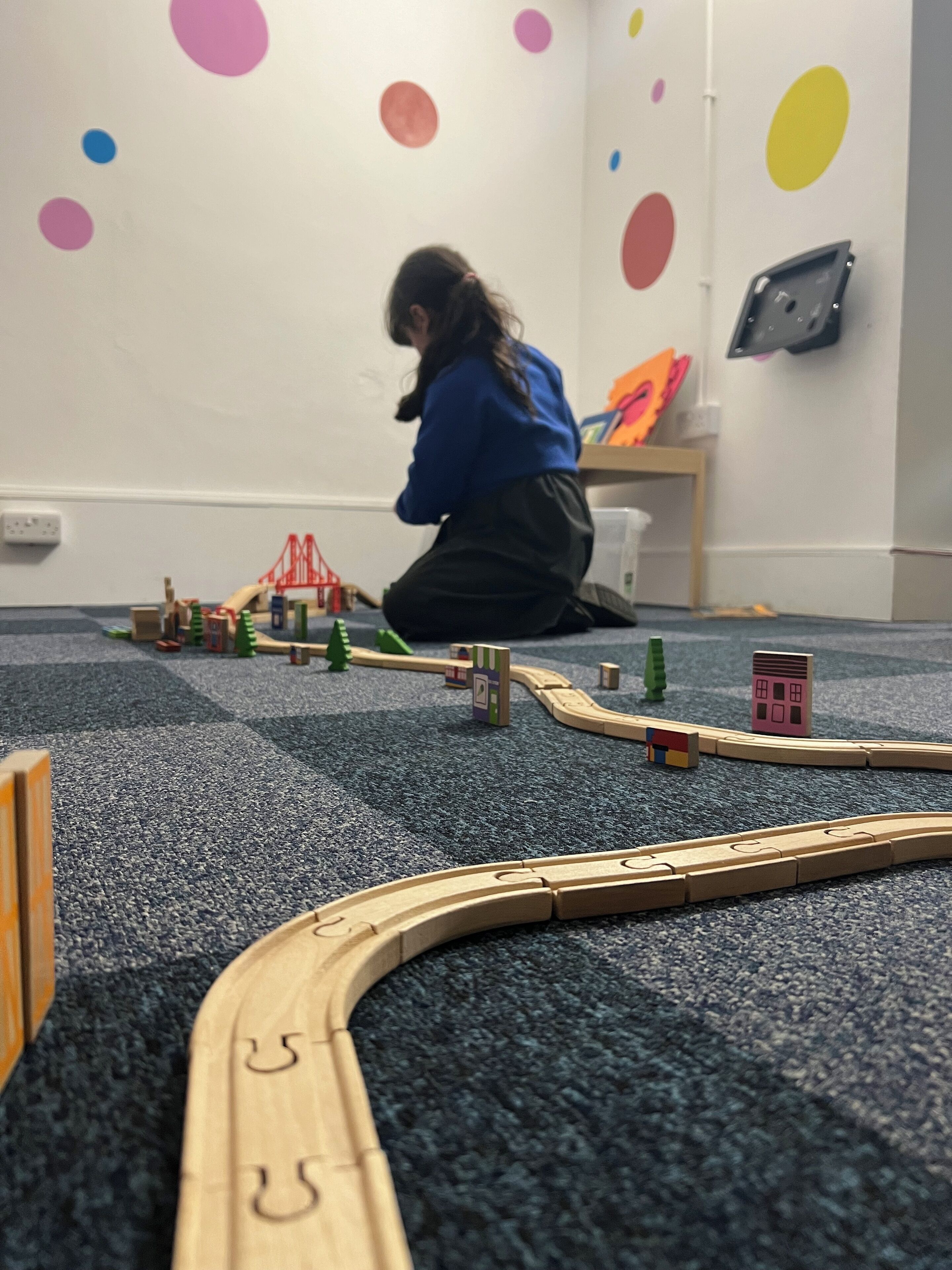 A child playing with wooden toy trains on a carpeted floor in a room with colorful wall decorations.