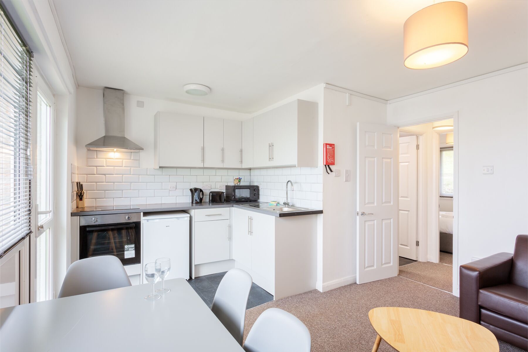 A compact kitchen with white cabinets, a microwave, and a sink. There is a dining area with a table and chairs, and a small wooden table.
