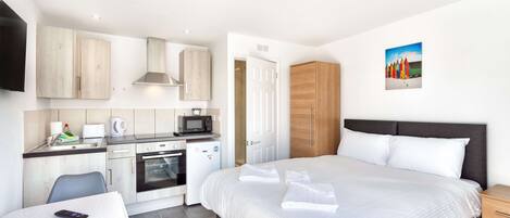 A modern hotel room with a kitchenette, a bed with neatly folded towels, a bedside table, and a painting on the wall.
