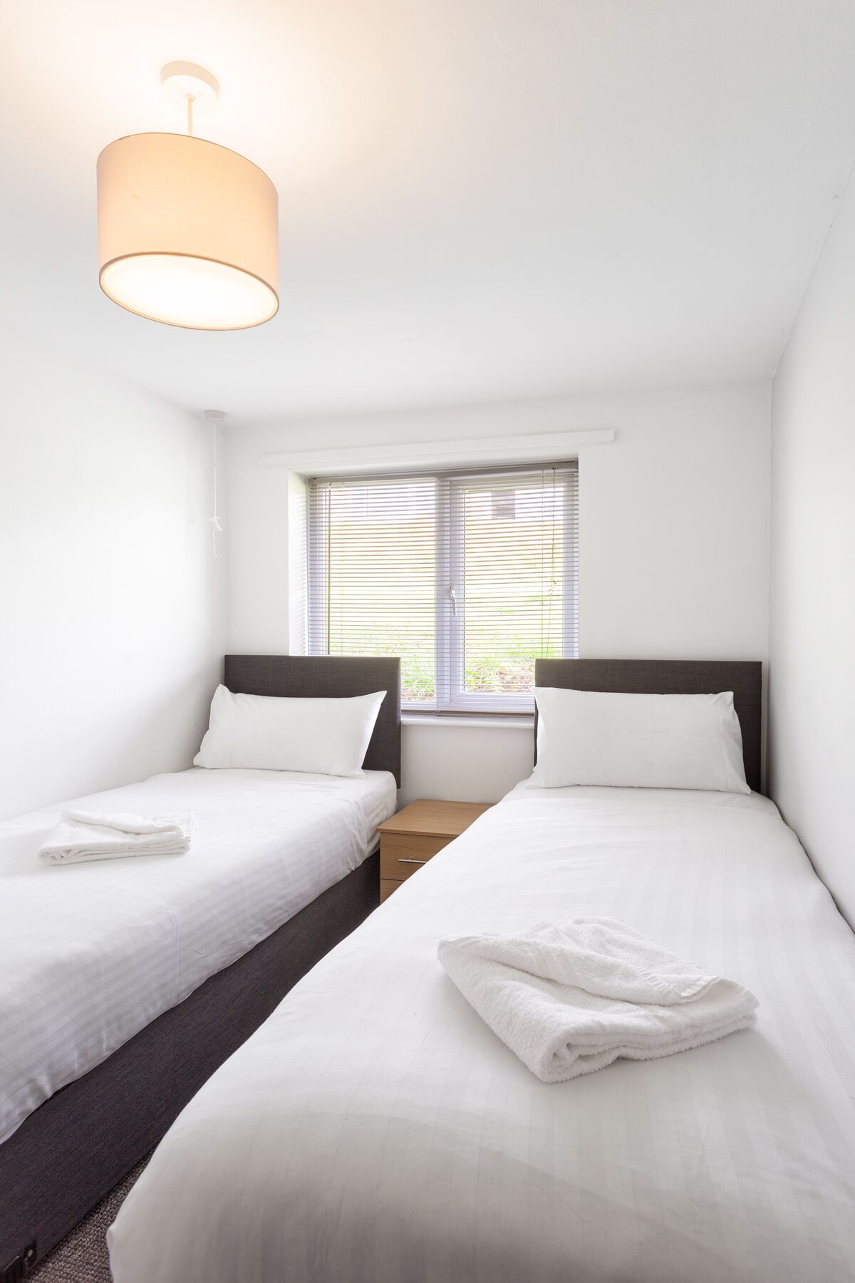 A hotel room with two single beds, a small window with blinds, a bedside table, and a hanging light fixture.