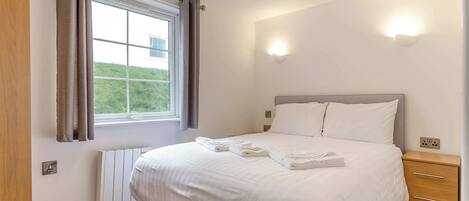 A bedroom with a bed, a radiator, a window with curtains, a bedside table, and a wall-mounted light.