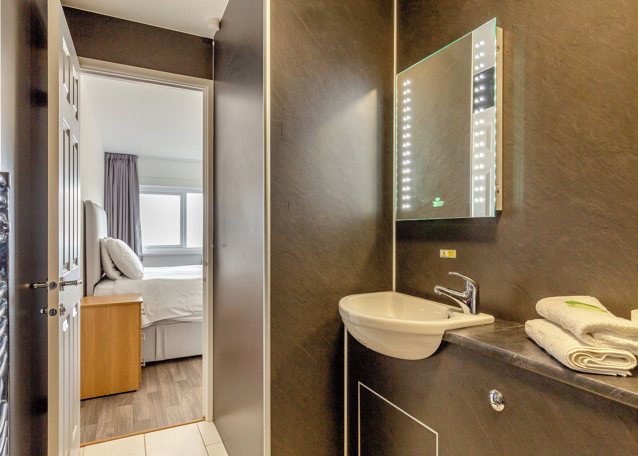 Un baño moderno con un espejo grande, un lavamanos y un inodoro. Se ve una mesita de noche de madera y una cama en la habitación contigua.