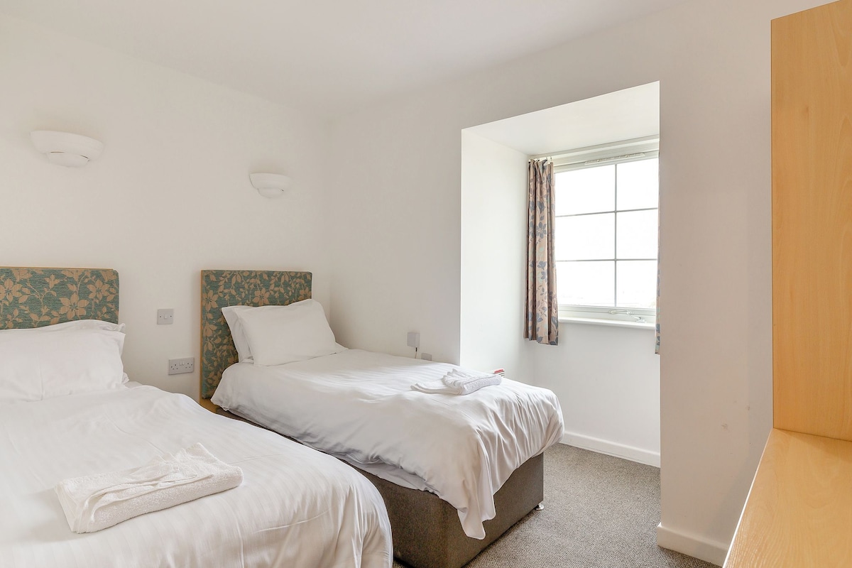 A clean, modern hotel room with two single beds, a large window with curtains, and a wooden wardrobe.