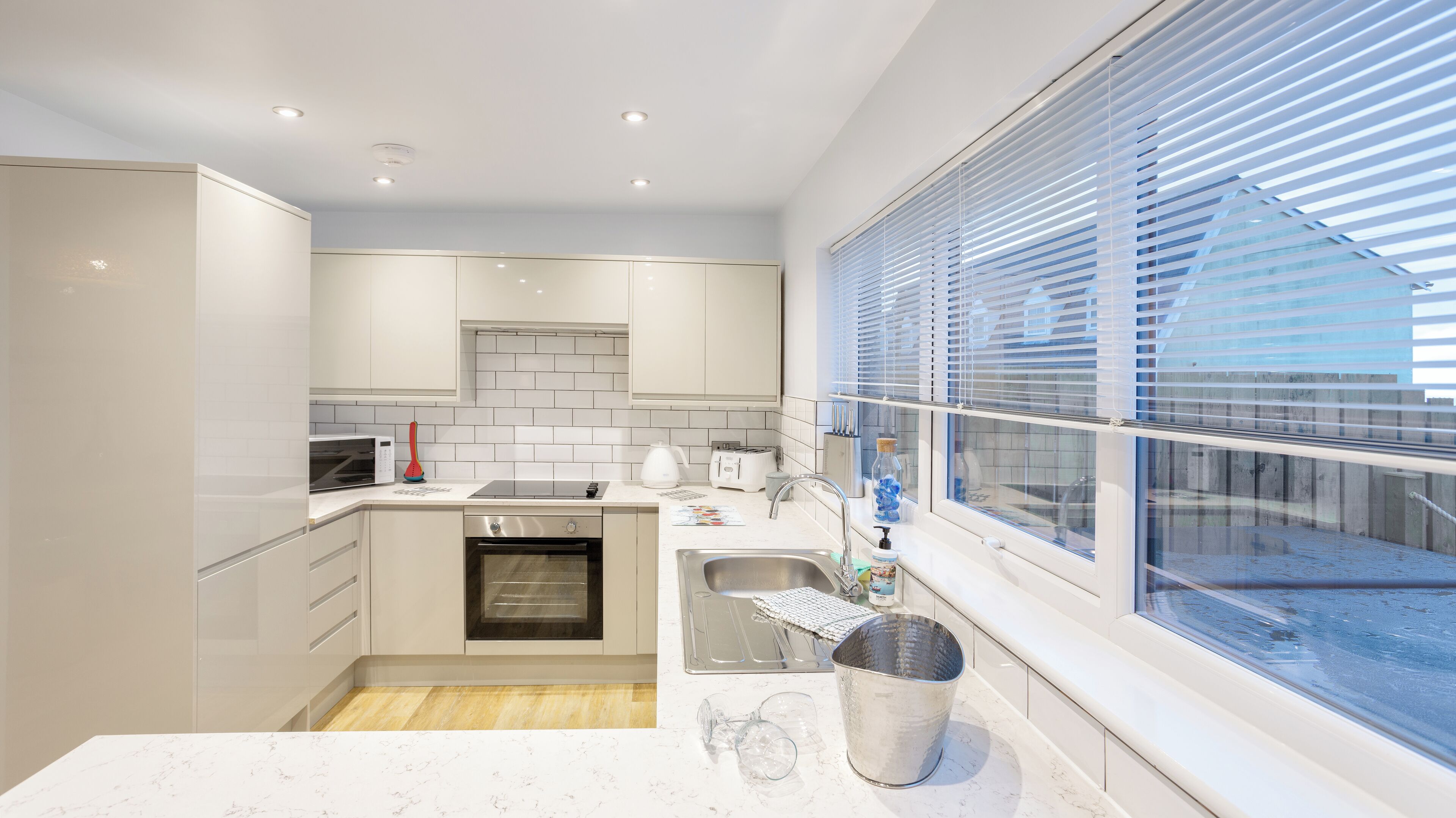 Una cocina moderna con armarios blancos, horno empotrado, lavamanos y ventana con persianas.