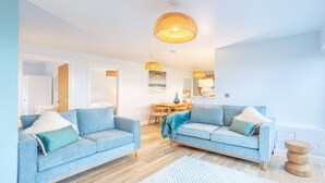 A modern living room with two light blue sofas, a wooden dining table, and a large woven pendant light. - Hot Tub Haven with Dreamy Sea Vista! (Ilfracombe)