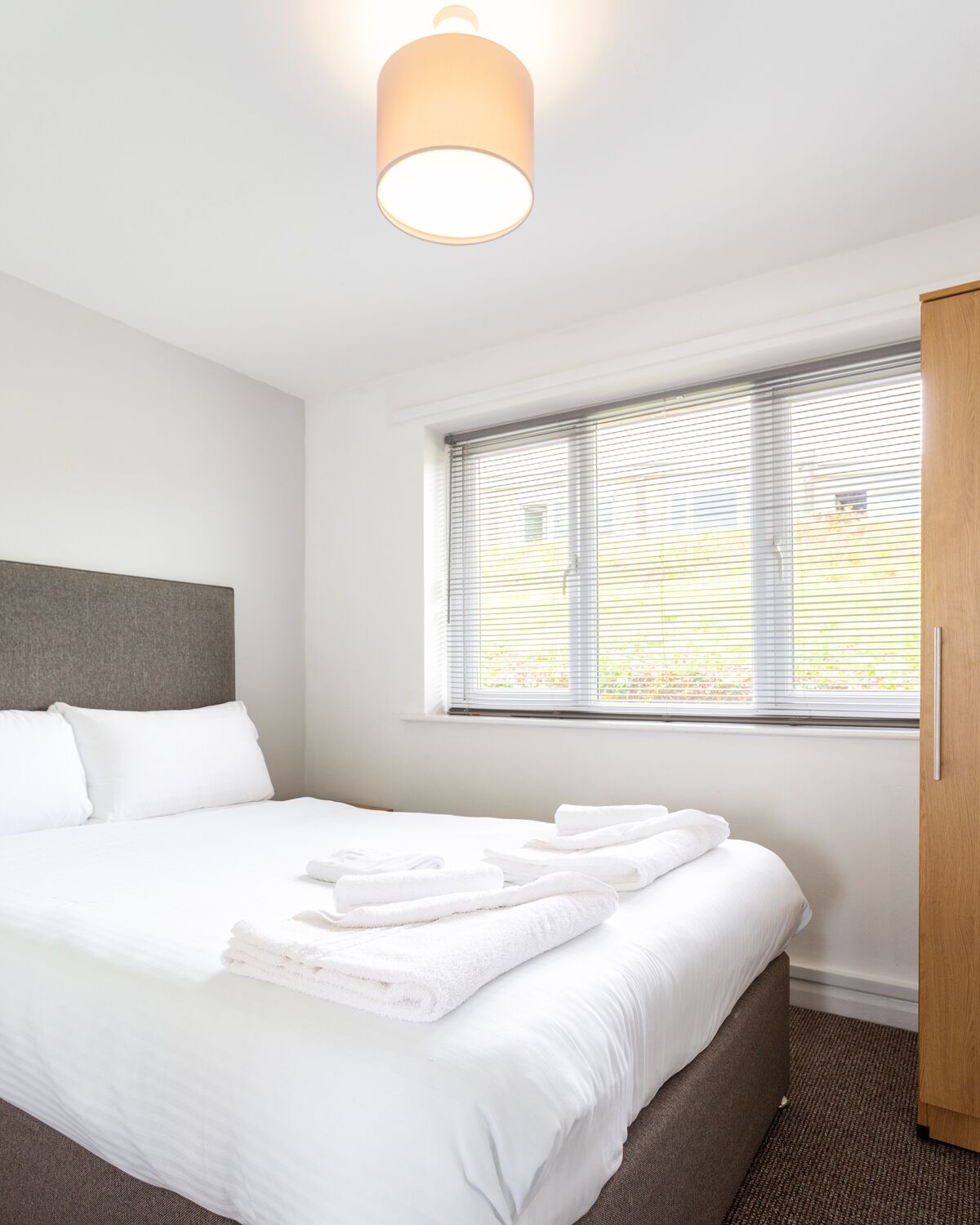 Una habitación de hotel moderna con una cama grande, cabecera gris, una ventana con persianas y un armario de madera.
