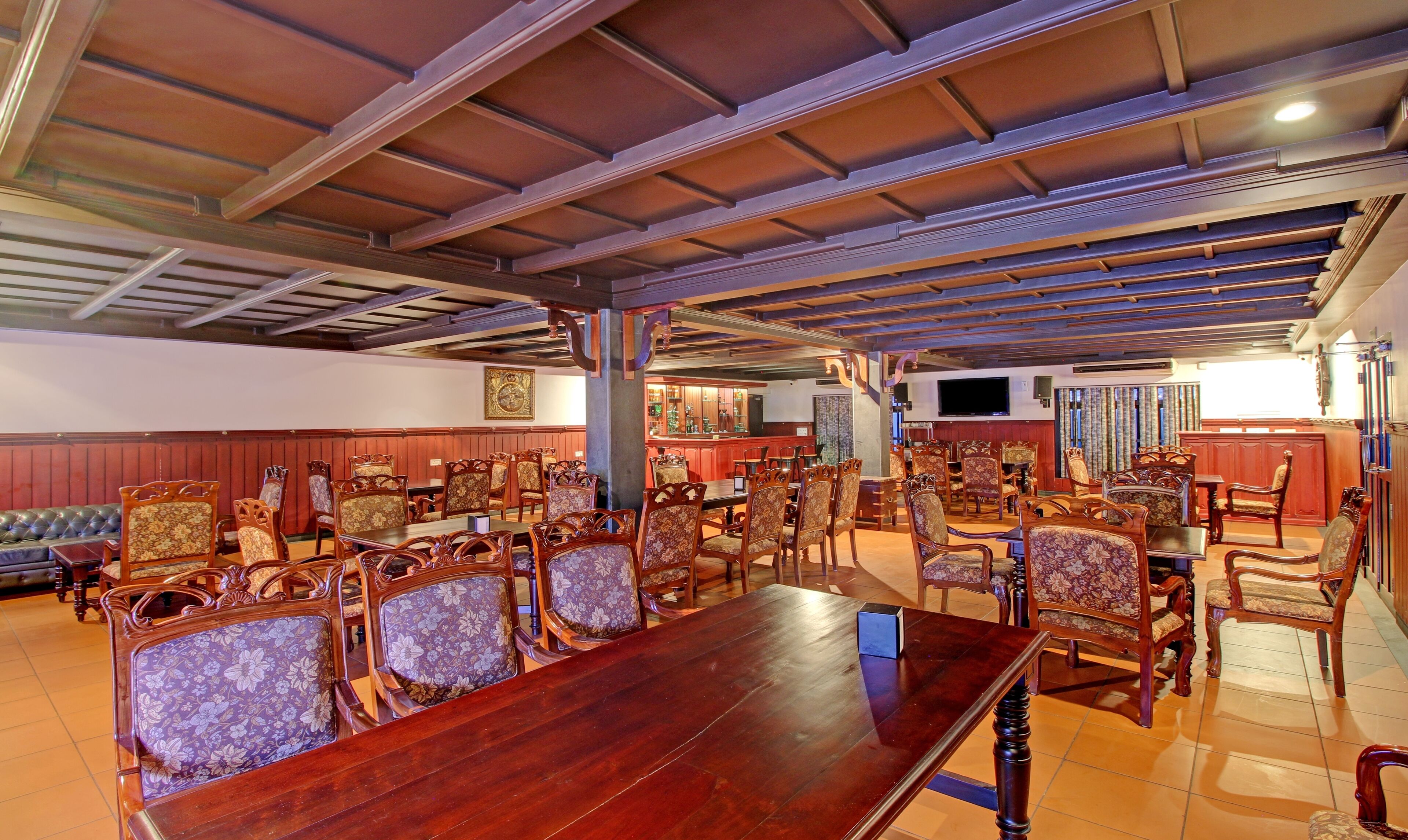 Una habitación amplia con una larga mesa de madera rodeada de sillas ornamentales, un televisor montado en la pared y una zona de bar al fondo.