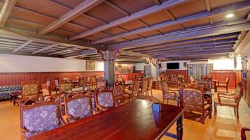 Una habitación amplia con una larga mesa de madera rodeada de sillas ornamentales, un televisor montado en la pared y una zona de bar al fondo.