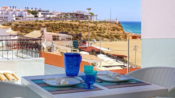 Un balcon avec une table dressée pour deux personnes, donnant sur une plage et une colline parsemée de bâtiments.