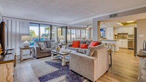 A spacious living room with a large window, a kitchen in the background, and a dining area. - Golf Cart ~ Seasonally Heated Pool ~ First Floor (Miramar Beach)