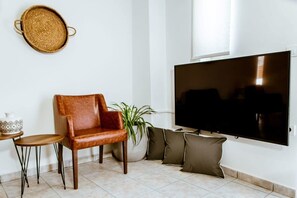 Uma sala de estar com uma poltrona de couro, uma mesinha redonda, um vaso de planta e uma grande televisão de tela plana fixada na parede.