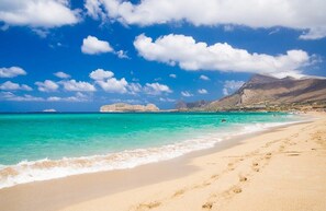 A beach with clear turquoise water, sandy shore, and distant rocky mountains under a blue sky with clouds.