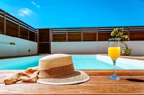 Zona all'aperto illuminata dal sole con piscina, tavolo in legno, cappello di paglia e bicchiere di succo d'arancia.