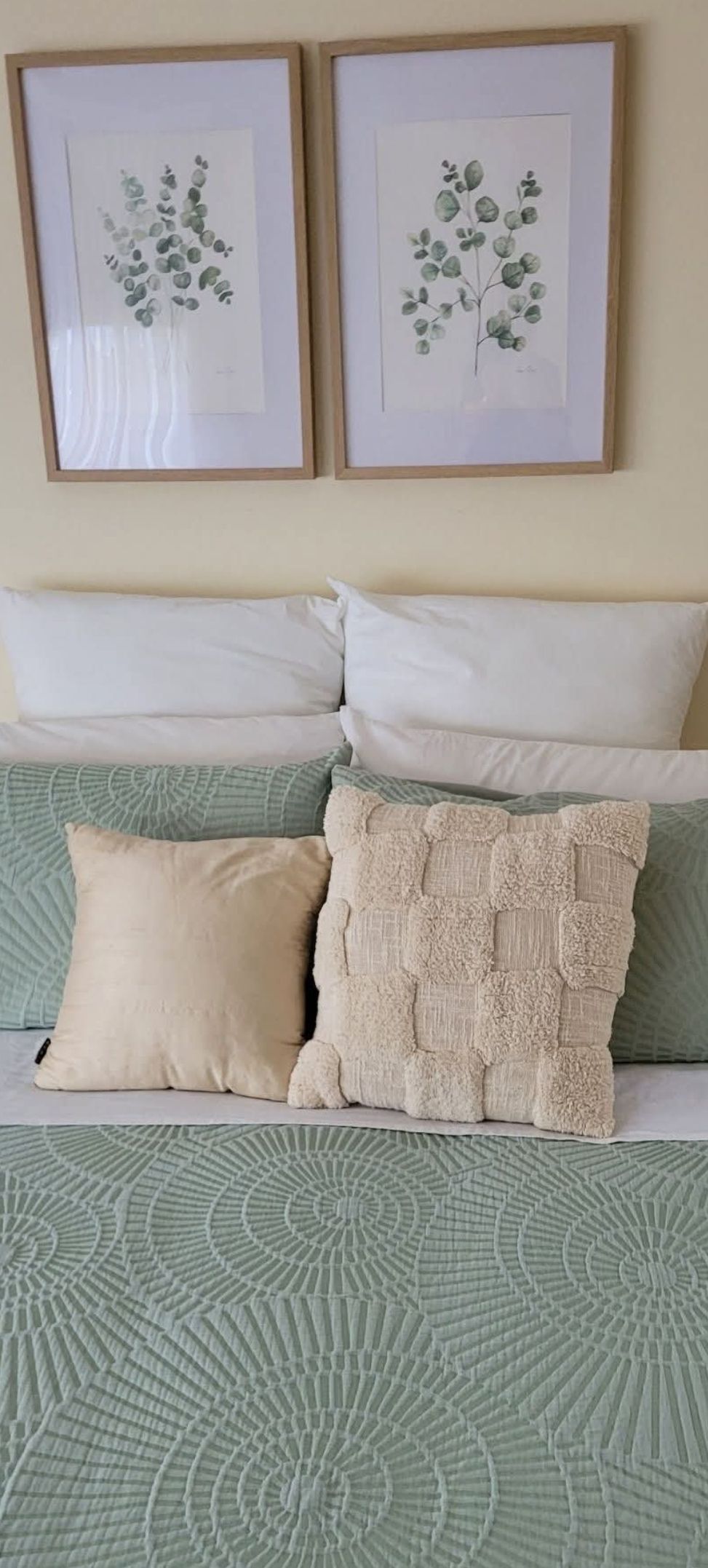 A bed with a green patterned bedspread, two framed botanical prints on the wall, and three pillows of different textures.