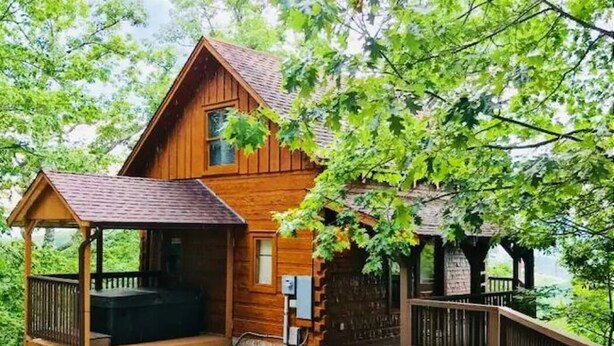 Una cabaña de madera con porche y jacuzzi, rodeada de árboles.